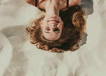 Close up top view of a smiling woman lying on beach sand. Close up of head and shoulders of a woman with closed eyes relaxing on beach. - JLPSF22553