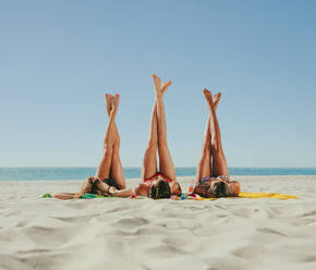 Drei Freundinnen liegen im Bikini am Strand und sonnen sich mit gekreuzten Beinen, die in den Himmel zeigen. Frauen entspannen am Strand an einem sonnigen Tag. - JLPSF22560