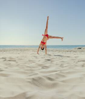 Woman in bikini doing an acrobatic move turning upside down on beach. Acrobatic woman upside down with both hands on ground and legs in air. - JLPSF22561