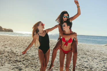 Drei Freundinnen in Bikini und Badeanzug, die in fröhlicher Stimmung am Strand spazieren gehen. Frau reitet huckepack auf ihrer Freundin, während sie sich am Strand vergnügt. - JLPSF22562