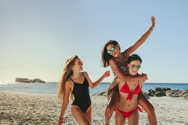 Three girlfriends in bikini and swimsuit walking on the beach in cheerful mood wearing sunglasses. Woman piggy riding on her friend while enjoying on the beach. - JLPSF22563