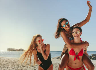 Three women in bikini and swimsuit walking on the beach in cheerful mood wearing sunglasses. Woman piggy riding on her friend while enjoying on the beach. - JLPSF22564