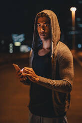 Fit junger Mann in Kapuzen-Shirt steht im Freien mit Handy. Fitness-Mann macht Pause nach dem Training im Freien bei Nacht. - JLPSF22569