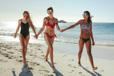 Drei Freundinnen im Bikini mit Sonnenbrille am Strand spazieren und halten sich an den Händen. Lächelnde Frauen im Urlaub Sonnenbaden am Strand zu Fuß mit Meer und Sonne Fackel im Hintergrund. - JLPSF22570