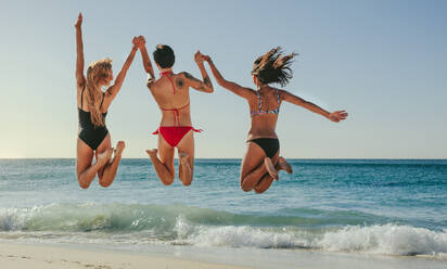 Rückansicht von drei Frauen im Bikini, die hoch springen, sich an den Händen halten und Spaß am Strand haben. Frauen im Urlaub, die sich am Strand vergnügen und in die Luft springen, mit Blick auf das Meer. - JLPSF22573