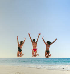 Rückansicht von drei Frauen im Bikini, die mit erhobenen Händen in die Luft springen und sich am Strand amüsieren. Frauen im Urlaub, die sich am Strand amüsieren und in die Luft springen, mit Blick auf das Meer. - JLPSF22574