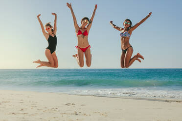 Drei Frauen im Bikini springen mit erhobenen Händen in die Luft und haben Spaß am Strand. - JLPSF22575