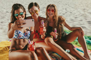 Drei Freundinnen im Bikini sitzen am Strand, halten Getränke und machen ein Selfie. Frauen im Urlaub am Strand posieren für ein Selfie mit Sonnenbrille. - JLPSF22576