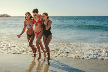 Drei Freundinnen im Bikini mit Sonnenbrille gehen am Strand und halten sich gegenseitig. Frauen im Urlaub Sonnenbaden am Strand gehen in den Gewässern mit Meer im Hintergrund. - JLPSF22582