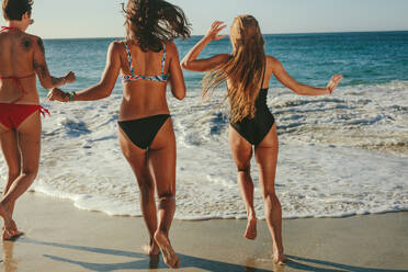 Rear view of three girlfriends in bikini walking on beach towards the sea. Women on vacation running towards the sea to take a dip. - JLPSF22583