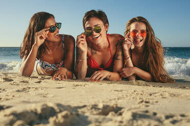 Drei Frauen im Bikini Sonnenbaden liegen auf der Vorderseite am Strand machen Gesichter halten ihre Sonnenbrille. Frau im Urlaub entspannen am Strand tragen Sonnenbrille Spaß mit Meer im Hintergrund. - JLPSF22586