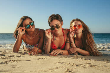 Drei Frauen im Bikini sonnenbaden liegend auf der Vorderseite am Strand halten ihre Sonnenbrille. Frau im Urlaub entspannen am Strand tragen Sonnenbrille Spaß mit Meer im Hintergrund. - JLPSF22587