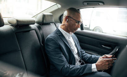 Business person checking cell phone sitting in his sedan on back seat. Man wearing earphones managing business work on the move sitting in car. - JLPSF22589