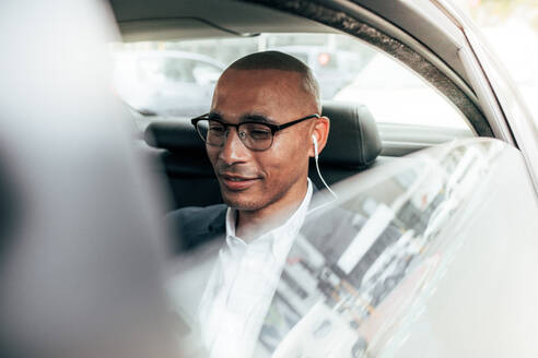 Business person wearing eyeglasses sitting in his sedan on back seat going to office. Man wearing earphones managing business work on the move sitting in car. - JLPSF22590