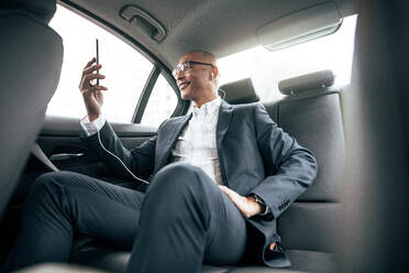 Geschäftsmann, der einen Videoanruf tätigt, indem er sein Handy hält, während er in seiner Limousine auf dem Rücksitz sitzt. Mann mit Kopfhörern, der unterwegs im Auto sitzend geschäftliche Arbeiten erledigt. - JLPSF22591