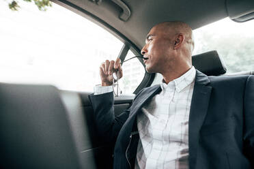 Businessman sitting in car and looking out of the window while commuting to office. Man holding eyeglasses sitting in his sedan on back seat. - JLPSF22592