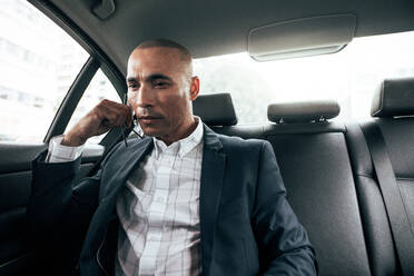 Businessman sitting in car looking down and thinking while commuting to office. Man holding eyeglasses in hand sitting in his sedan on back seat. - JLPSF22594