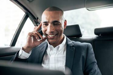 Ein Geschäftsmann arbeitet an einem Laptop, während er in seiner Limousine ins Büro fährt. Ein lächelnder Mann, der eine Brille in der Hand hält, verwaltet geschäftliche Aufgaben, während er im Auto sitzt. - JLPSF22597