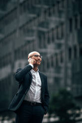 Lächelnder Geschäftsmann im Gespräch über Handy im Freien stehend. Mann in formeller Kleidung, der auf der Straße geht und mit dem Handy spricht. - JLPSF22599