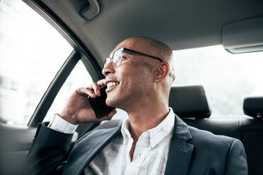Smiling businessman wearing eyeglasses talking over cell phone sitting in his sedan looking out the window. Man managing business on mobile phone sitting in car. - JLPSF22601