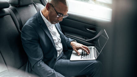 Geschäftsmann, der an einem Laptop arbeitet, während er in seiner Limousine auf dem Rücksitz ins Büro fährt. Unternehmer, der unterwegs im Auto sitzend geschäftliche Arbeiten erledigt. - JLPSF22602