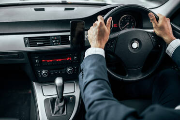 Rear view of man in formal clothes holding the steering wheel of his car with both hands. Businessman driving car with mobile phone clipped to dashboard. - JLPSF22604