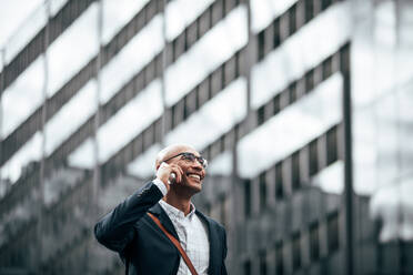 Smiling businessman talking over cell phone while commuting to office with a glass facade building in background. Man in formal clothes carrying office bag walking on street while talking on mobile phone. - JLPSF22609