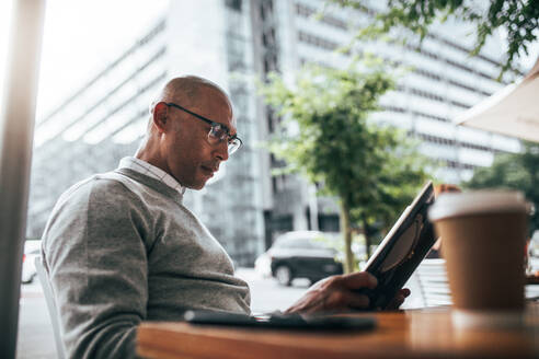 Businessman in sweater reading a book sitting at a restaurant with coffee cup and mobile phone on the table. Man in eyeglasses sitting in a street side restaurant reading book. - JLPSF22614