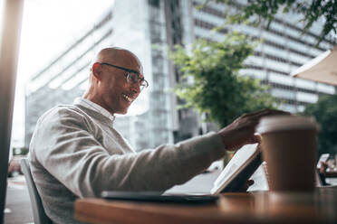 Lächelnder Geschäftsmann im Pullover, der in einem Restaurant mit Kaffeetasse und Handy auf dem Tisch sitzt und ein Buch liest. Mann mit Brille, der in einem Restaurant am Straßenrand sitzt und ein Buch liest. - JLPSF22616