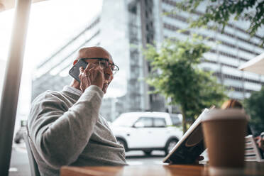 Geschäftsmann im Pullover im Gespräch auf dem Handy beim Lesen eines Buches sitzen in einem Restaurant mit Kaffeetasse auf dem Tisch. Mann mit Brille sitzen in einer Straße Seite Restaurant im Gespräch auf dem Handy und ein Buch zu lesen. - JLPSF22617