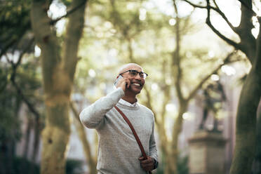 Smiling businessman talking over cell phone while walking on city street to work. Man walking through park wearing office bag talking on mobile phone. - JLPSF22625