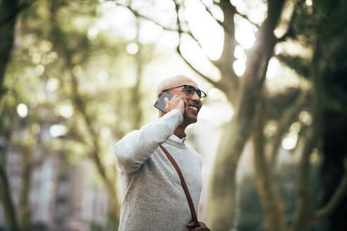 Mann, der durch einen Park geht und eine Bürotasche trägt und mit einem Mobiltelefon spricht. Lächelnder Geschäftsmann, der über ein Mobiltelefon spricht, während er auf einer Straße in der Stadt zur Arbeit geht. - JLPSF22626