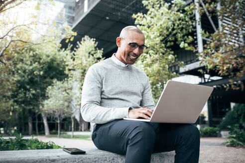 Lächelnder Geschäftsmann, der im Freien sitzend an einem Laptop arbeitet, mit einem Mobiltelefon an seiner Seite, Unternehmer, der im Freien sitzend seine Arbeit erledigt. - JLPSF22631