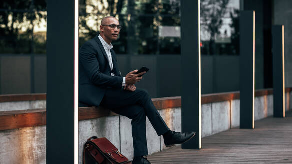 Unternehmer, der am Straßenrand sitzt und sein Mobiltelefon und seine Bürotasche in der Hand hält. Geschäftsmann, der unterwegs im Freien sitzt und seine Geschäfte führt. - JLPSF22633