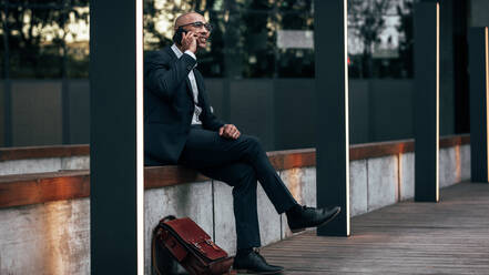 Entrepreneur sitting on the side of a street talking on mobile phone with his office bag by his side. Businessman managing business on the move sitting outdoors. - JLPSF22634