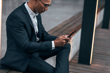 Unternehmer tippt Text in Handy sitzen auf Seite Geländer im Freien. Geschäftsmann sitzt im Freien verwalten Arbeit mit Handy. - JLPSF22635