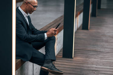 Unternehmer in formeller Kleidung sitzt auf einem Geländer im Freien und tippt Text in sein Handy. Geschäftsmann sitzt im Freien und verwaltet seine Arbeit mit seinem Handy. - JLPSF22636