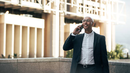 Smiling businessman talking over cell phone standing on terrace of a building. Man in formal clothes standing on a building talking on mobile phone. - JLPSF22637