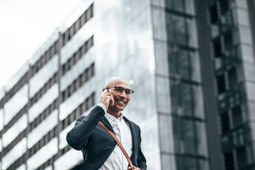 Smiling businessman talking over cell phone while commuting to office with a glass facade building in background. Man in formal clothes carrying office bag walking on street while talking on mobile phone. - JLPSF22638
