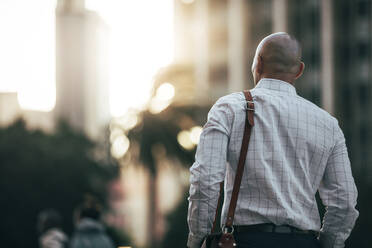Rückansicht eines Mannes in formeller Kleidung, der auf der Straße zum Büro geht und eine Bürotasche trägt, mit einer Sonnenfackel im Hintergrund. Ein Mann, der zu Fuß ins Büro geht. - JLPSF22639