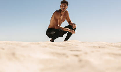Man sitting on his toes and relaxing during workout at the beach. Bare chested man sitting at the beach in fitness gear during workout. - JLPSF22642
