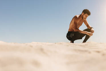 Mann sitzt auf den Zehen und entspannt sich beim Training am Strand. Nackter Mann sitzt am Strand in Fitnesskleidung und berührt Sand beim Training. - JLPSF22643