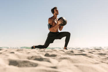 Ein Mann macht ein Fitnesstraining am Strand mit einem Medizinball. Ein athletischer Mann mit nacktem Oberkörper macht Dehnübungen mit einem Medizinball und trägt drahtlose Kopfhörer und ein Mobiltelefon an der Armbinde. - JLPSF22646