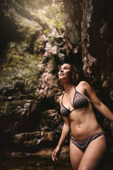 Woman wearing bikini looking up at something interesting while standing inside a pond. Female admiring the view from a pond in jungle. - JLPSF22650