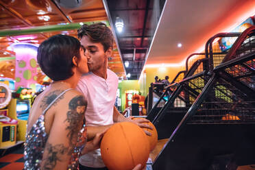 Paar, das sich in einer Spielhalle küsst und Basketbälle in der Hand hält. Mann und Frau in romantischer Stimmung in einer Spielhalle, die Spaß am Spielen haben und sich küssen. - JLPSF22656