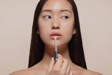 Portrait of beautiful young woman applying transparent lip gloss with applicator. Asian female model looking away while doing make up against beige background. - JLPSF22665