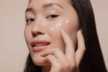Nahaufnahme einer jungen Frau, die eine Feuchtigkeitscreme auf ihr Gesicht aufträgt. Eine asiatische Frau sieht glücklich aus, während sie ihre Hautpflege betreibt. - JLPSF22667