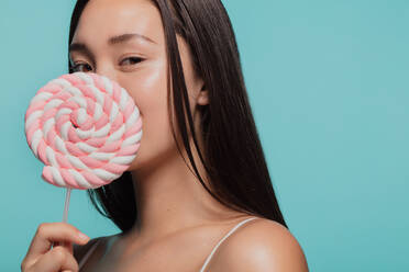 Porträt einer jungen Frau mit einem Bonbon. Asiatisches Mädchen hält einen großen Swirl-Lutscher vor ihrem Gesicht vor blauem Hintergrund. - JLPSF22669