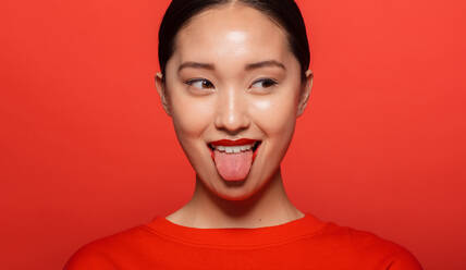 Close up of young asian woman sticking out tongue and looking away. Korean female model making funny face against red background. - JLPSF22678