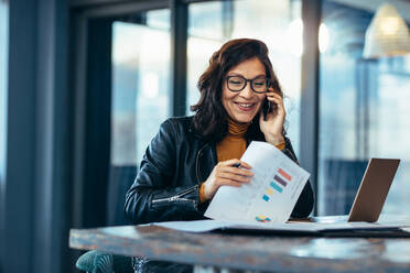 Glückliche asiatische Frau bei der Arbeit im Büro, die am Telefon spricht, während sie Papierkram am Arbeitsplatz erledigt. - JLPSF22701